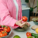 Person in pink overshirt holding a fresh tomato with various organic vegetables on a countertop.
